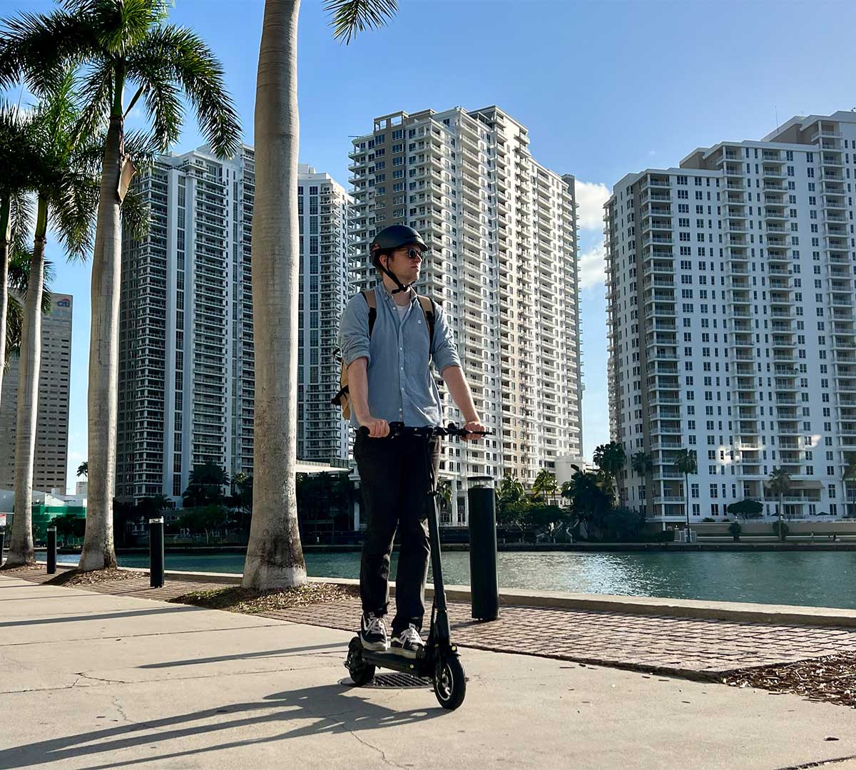 Man on an electric scooter by the waterfront with cityscape in the background.