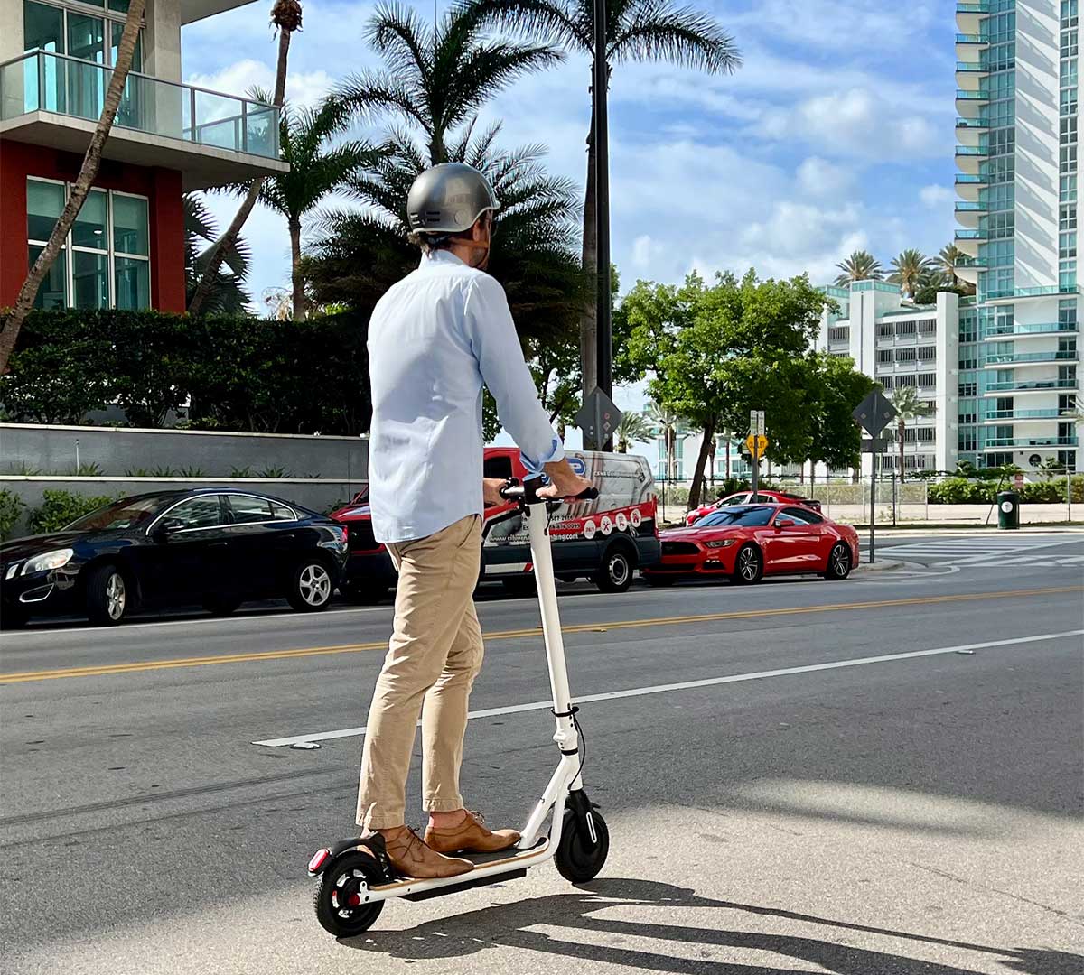 An individual in business casual attire and wearing a helmet responsibly rides an electric scooter alongside a city street, highlighting the importance of road safety and the shared use of streets between electric scooters and traditional vehicles.