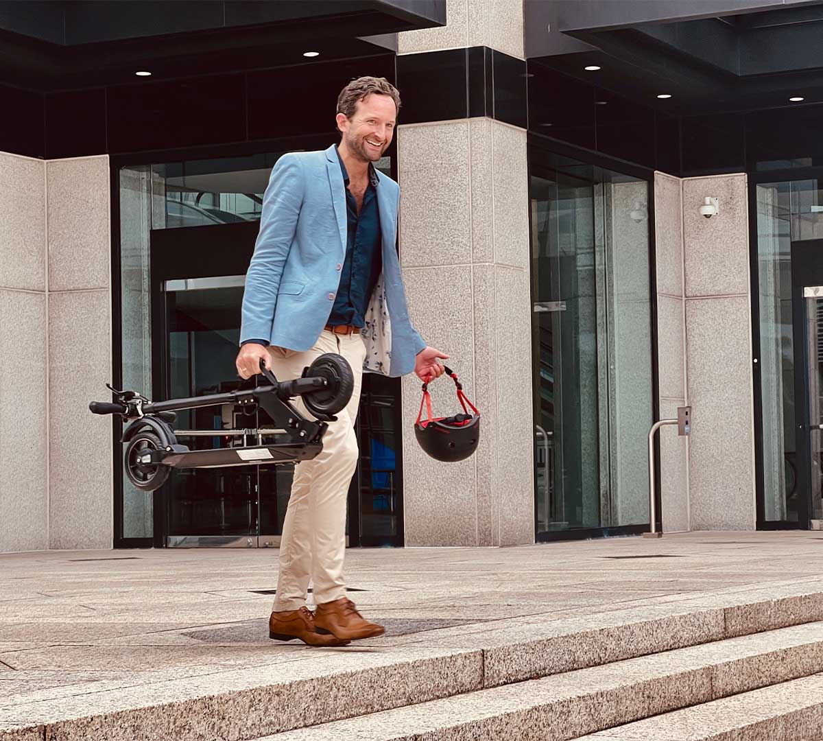 A stylish man in business casual attire exits a modern building carrying a folded electric scooter, representing the synergy of electric scooter laws with urban lifestyle.