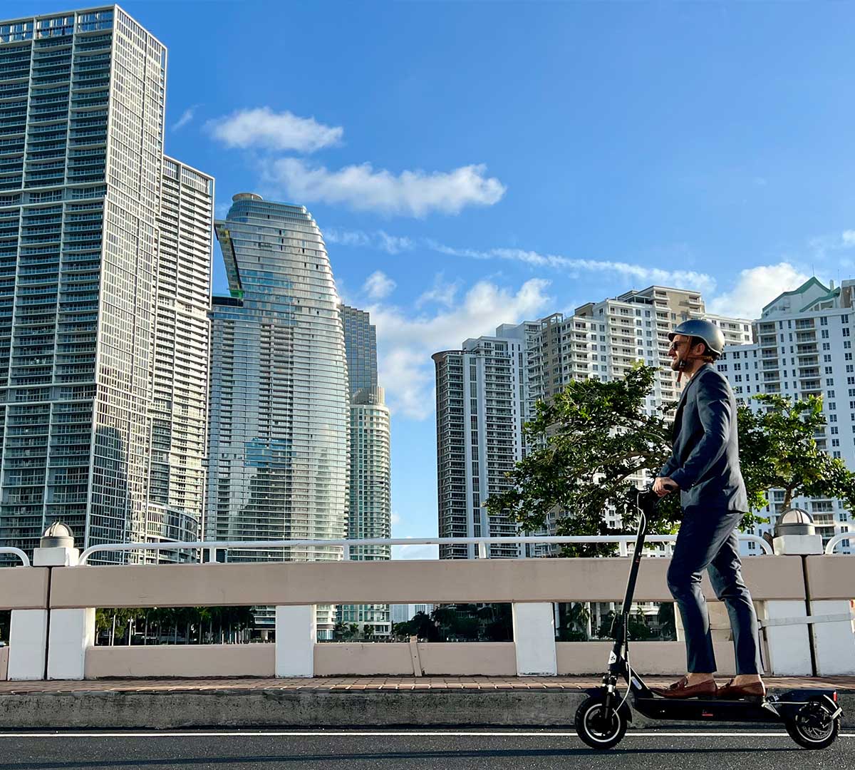 A businessman riding an electric scooter across a bridge with a backdrop of modern skyscrapers, symbolizing the integration of eco-friendly transportation in urban settings.