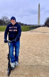 Smiling man named Tucker wearing a Harley Davidson hoodie and sunglasses standing with a Horizon and Mosquito electric scooter in front of the Washington Monument, symbolizing the freedom and joy of urban e-scooting.
