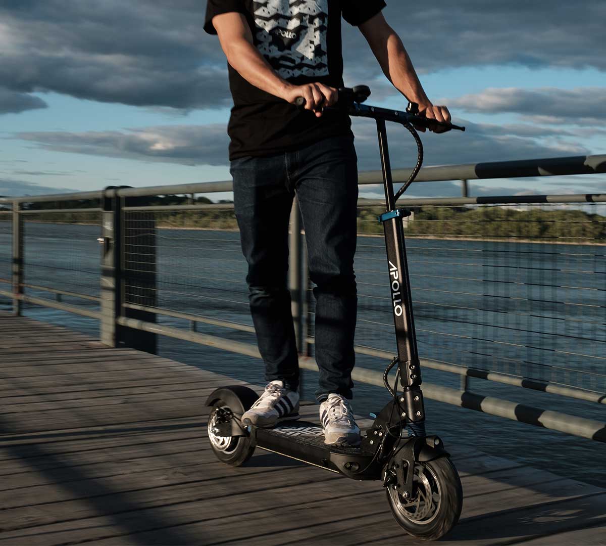 A person rides a high-quality electric scooter branded 'APOLLO' along a riverside boardwalk, demonstrating the scooter's sturdy design and adherence to safety standards with its robust frame and well-engineered wheels, ensuring a safe commuting experience