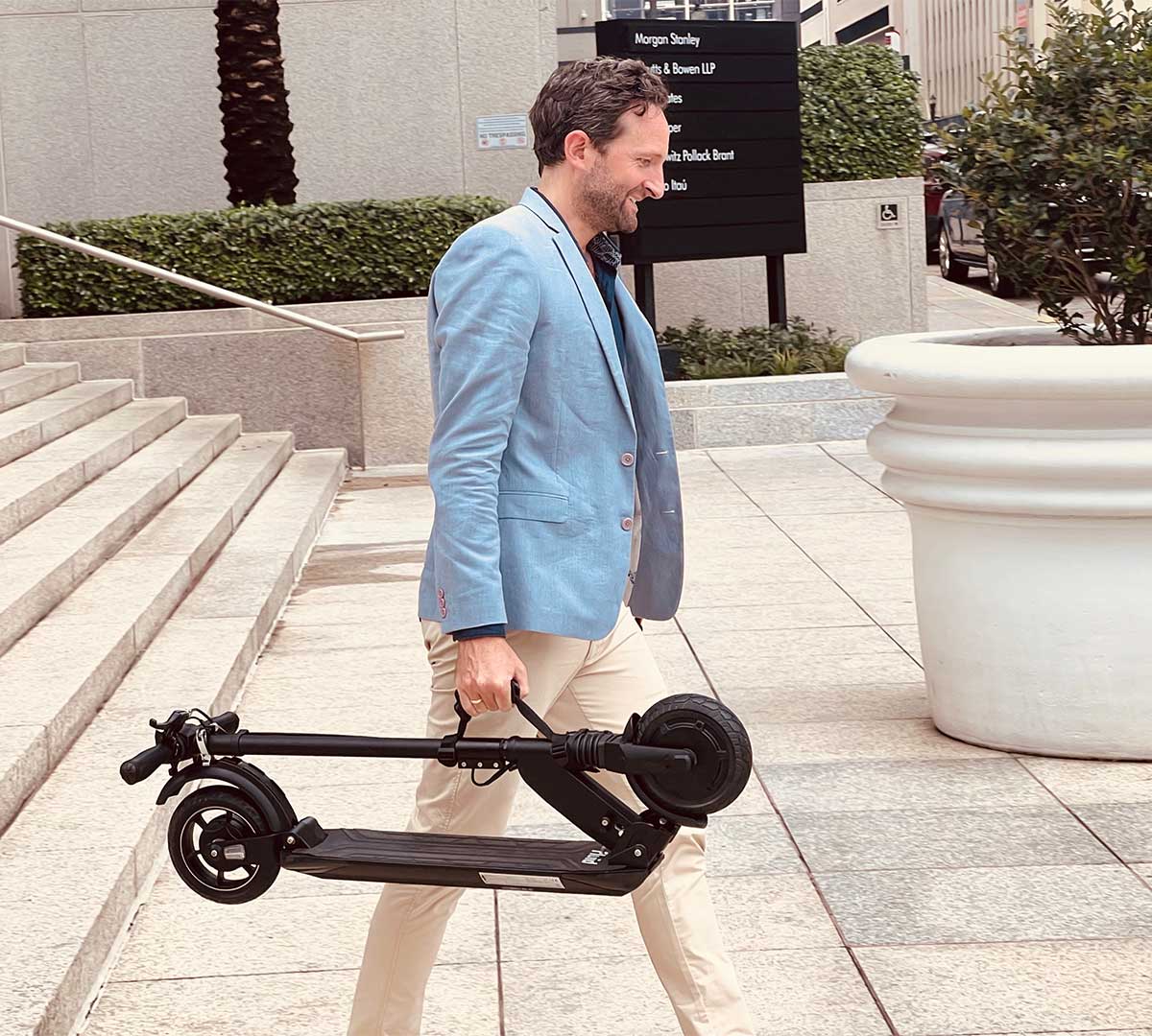 An adult male in a light blue blazer and beige trousers happily carries a small, foldable black electric scooter, ideal for city commuting, as he walks by a business complex.