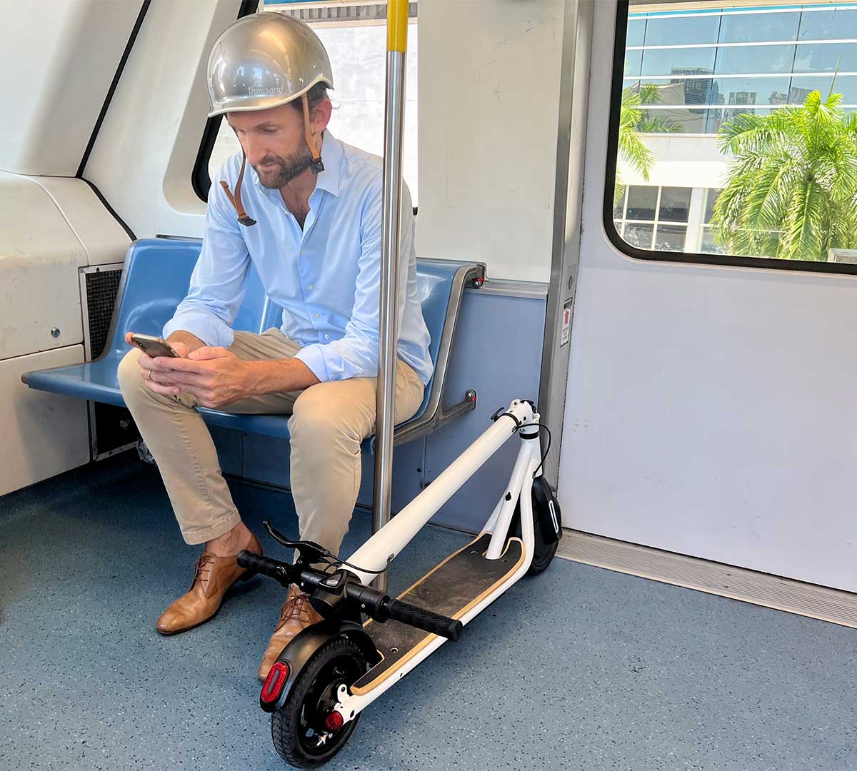 A commuter in smart casual attire sits on public transport, engrossed in his smartphone, with a portable electric scooter by his side, showcasing the innovative integration of personal electric vehicles in daily transit.