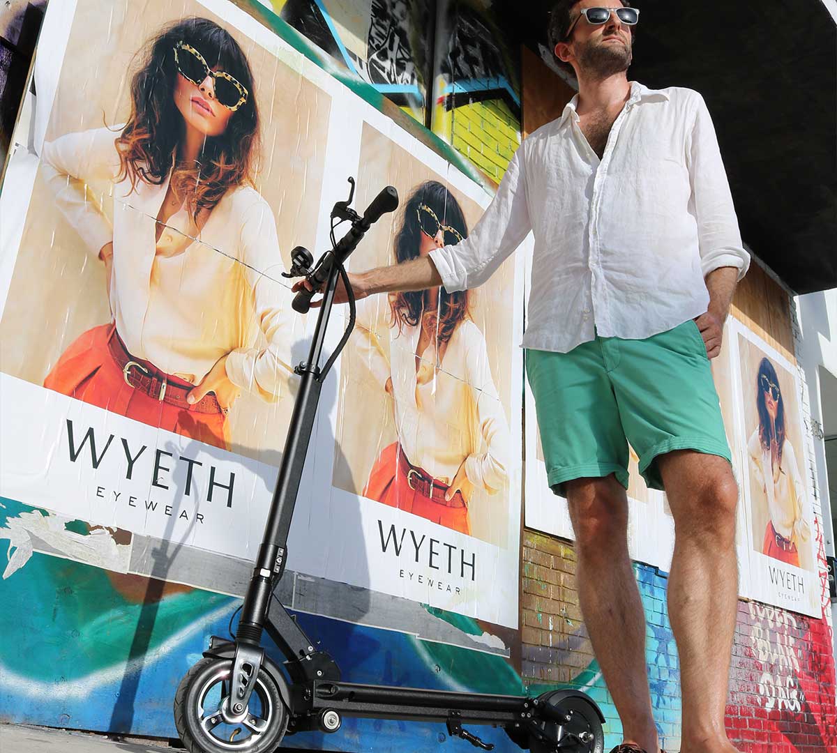 Man standing confidently beside a Varla Electric Scooter under a vibrant street art billboard, embodying the urban lifestyle highlighted in 'The Ultimate Guide to Varla Electric Scooters'.